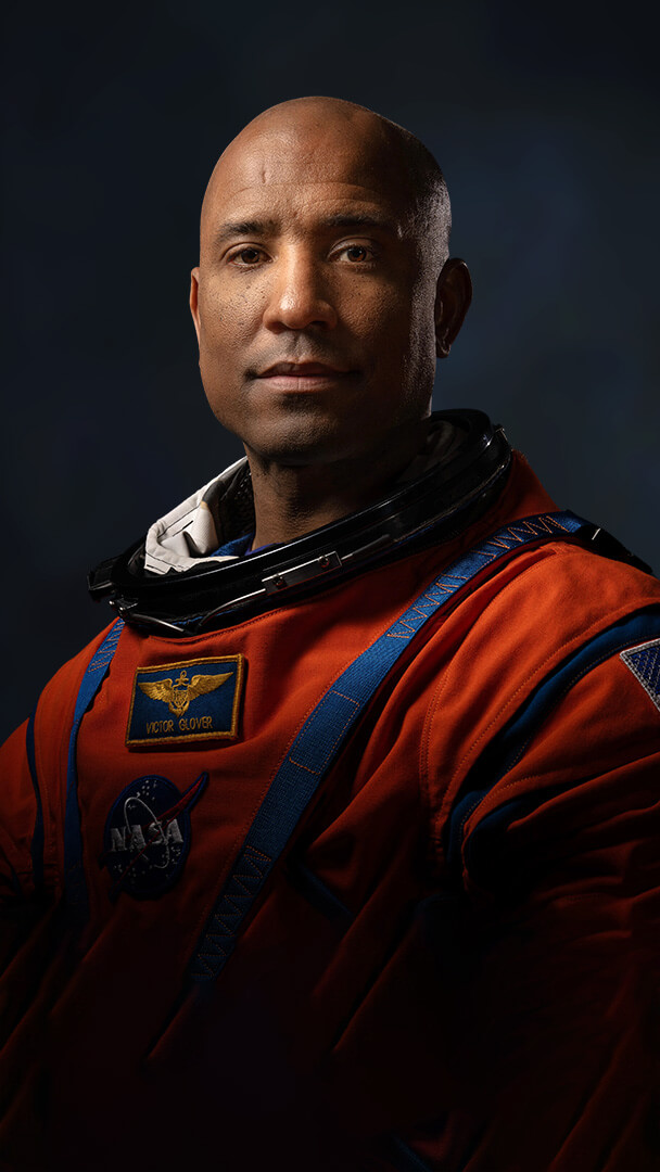 Official Nasa portrait of Victor Glover in his orange spacesuit showing him from mid-waist. The high contrast lighting from the side makes him look heroic. He is looking directly at the camera with a stoic expression.