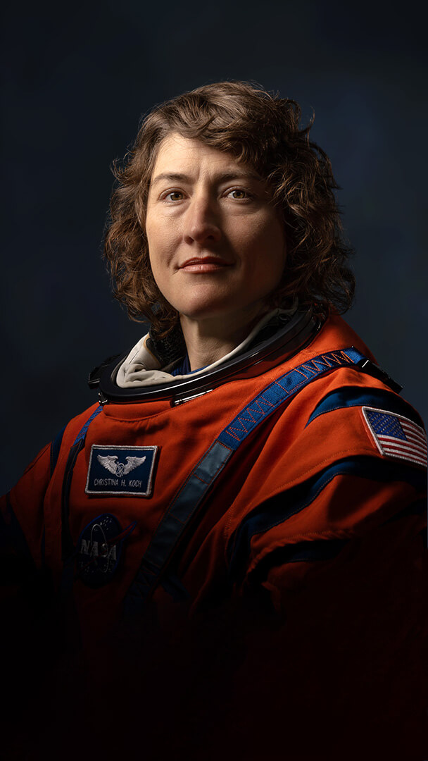 Official Nasa portrait of Christina Koch in her orange spacesuit showing her from mid-waist. The lighting is high contrast and dramatic making him look heroic. Her hair falls in curls either side of her face. She stares towars the camera looking both contemplative and determined.