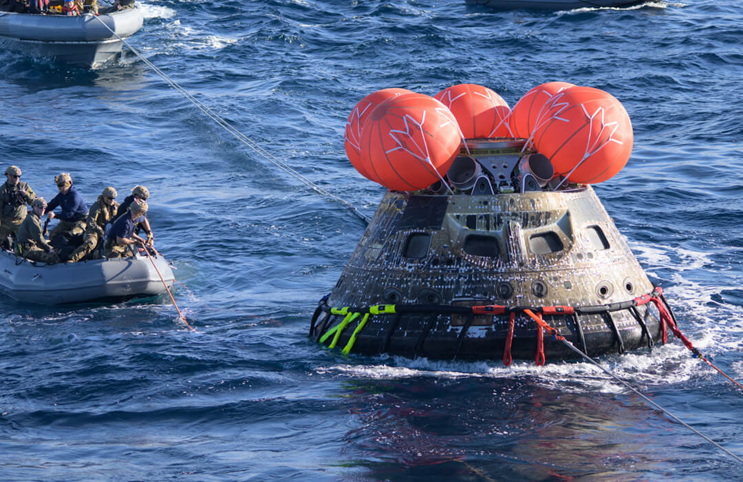 The Orion capsule from Artemis I being recovered from the ocean by the US Navy. The capsule is floating in the ocean with five bright orange ballons gathered around the top. The outer surface of the capsule looks burnt from the heat of reentry. An inflatable dingy with six men in military fatigues and helmets floats to the left with a line in the water. Another larger dingy, almost out of shot in the background, already has a line attached to the capsule. Two more lines attached to the front of the capsule are pulled taut and lead out of the frame. We can’t see what they are attached to but it appears to be slowly towing the capsule though the water.