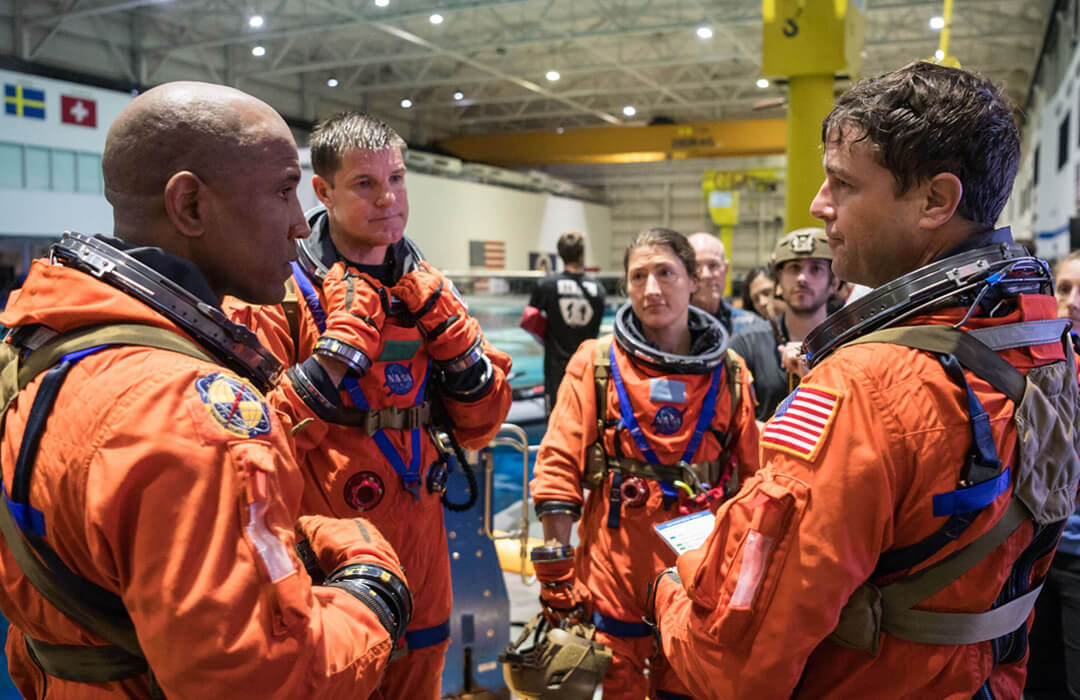 The four Artemis II astronauts in their orange spacesuits stand together in a circle next to an indoor pool. Victor Glover and Reid Wiseman look at each other in the foreground with Jeremy Hansen and Christina Koch behind them. They look as if they are having a debrief after some aspect of training has maybe not gone quite as expected. Other people stand in the background, looking on but not part of the discussion between the astronauts.
