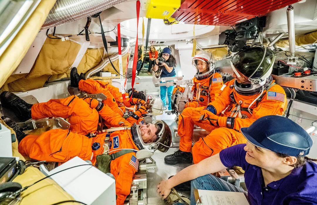 A group of Nasa engineers in bright orange spacesuits sit in the Orion crew capsule. The inside of the capsule looks cramped with every surface covered in equipment or silver ducts. An engineer in a spacesuit in the foreground smiles as he tries to either lie down on, or get up from, one of the seats. Near him in the extreme foreground a woman in a blue polo shirt and blue cap also smiles as she looks at him struggling to move. A photographer, also in blue polo shirt and blue hat, stands in the background holding her camera ready to take a shot. She is framed by the open hatch. Further behind her a blonde woman in a red top is sitting outside with her hands on her knees, smiling at the slightly chaotic scene taking place in the capsule.