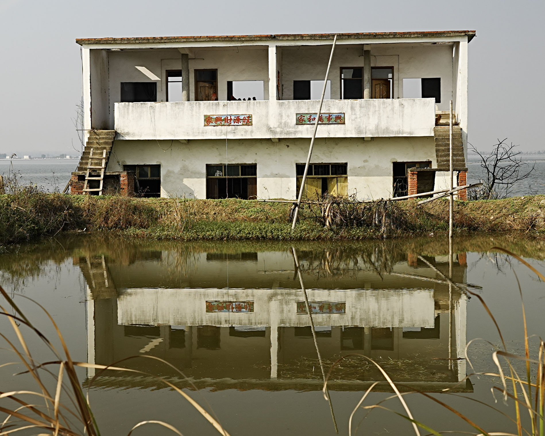 一棟被遺棄的兩層白色建築矗立在靜止的水塘旁,斑駁的外牆倒映在水面上。建築的窗戶與門已缺失,油漆剝落,兩側設有外露樓梯。