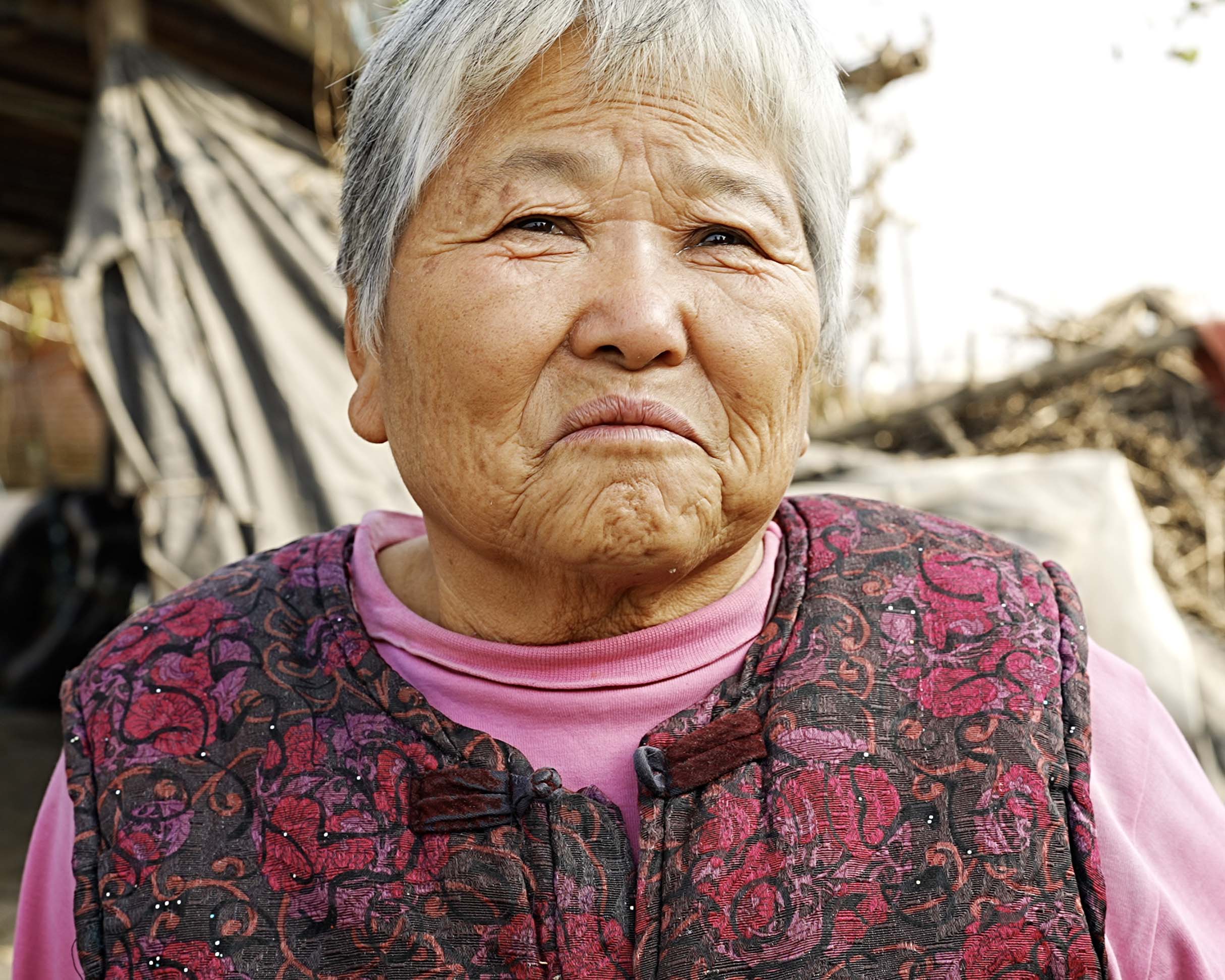 郭太太身穿粉紅色上衣與紅黑花紋的深色絎縫背心,站在戶外,身後可見帆布棚、木製結構與乾枯的植被。