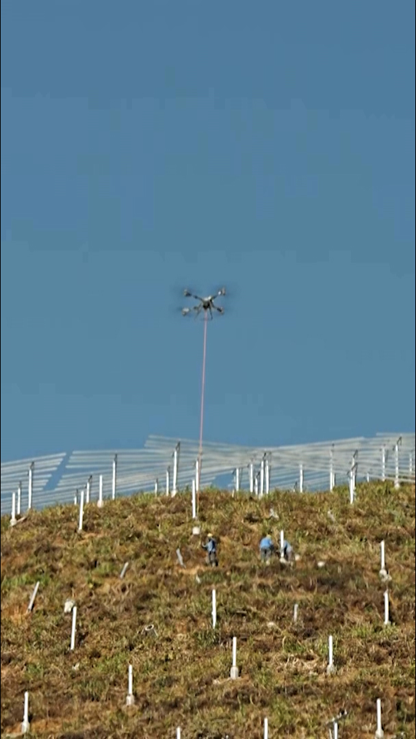 一架無人機懸停在山坡上空,坡面插著整齊排列的白色標記柱。身穿藍色工作服、戴著黃色安全帽的工人分布在山坡各處,山頂沿線可見金屬欄杆,在晴朗藍天下格外醒目。
