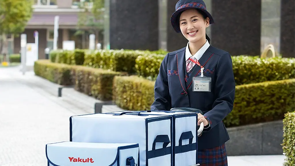A Yakult delivery worker in a navy uniform hands a tray of drinks to a smiling woman at the doorway of a wooden house in Japan (Credit: Yakult Honsha)