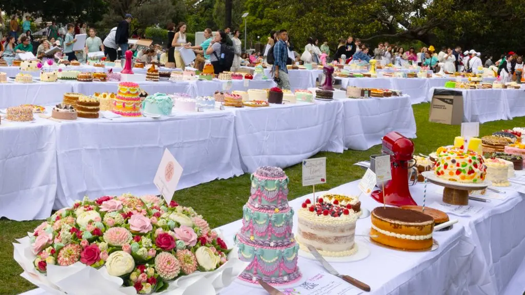 A woman with turquoise hair and glasses, green earrings and necklace and a bright turquoise top, with green and turquoise bangles, holding a carrot cake