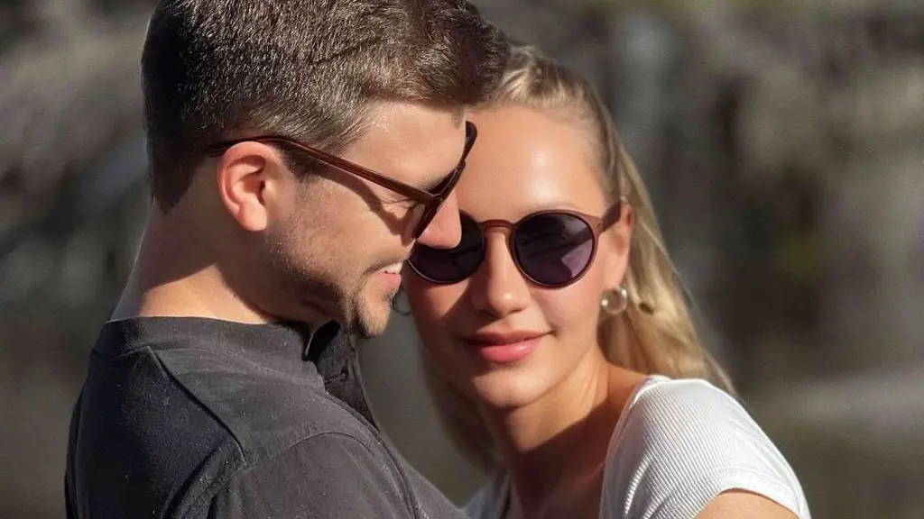 Two people standing close together in the foreground, wearing padded winter jackets, with one in a dark green coat and the other in a white coat and knitted headband. Snow is visible on the clothing. Behind them are snow-covered trees and branches.