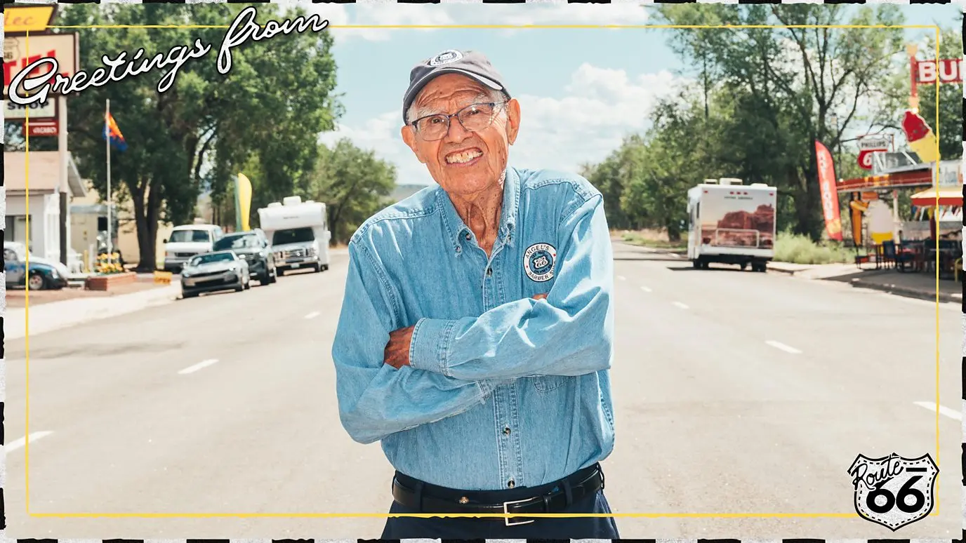 Angel Delgadillo stands in the middle of wide street on Route 66 in a postcard-style image reading "Greetings from Route 66" (Credit: Johnny Kompar/ Serenity Strull/ BBC)