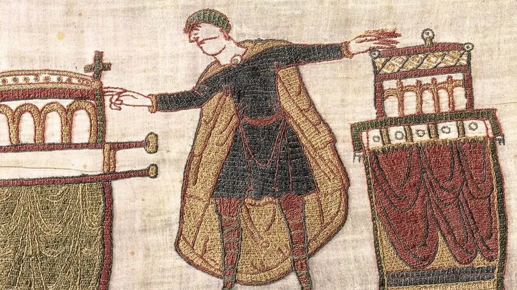 A woman with a white coat, visor and iPad looks closely at a scene from the Bayeux Tapestry