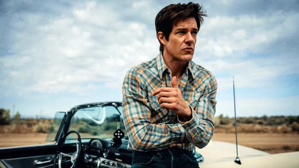 Brandon Flowers holds a stetson in his right hand and gives a wry look to the camera as he poses outside a stereotypical American motel