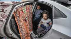 Two young Palestinian children shelter in a car with carpets and rugs hung over the windows to create a make-shift shelter