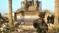An IDF soldier walks in front of frame as an enormous bulldozer clears rubble behind him