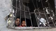 A woman and child look out a barred window. Chicken wire that appears to have covered the window is in tatters