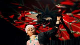 A boy spins a large, multi-coloured skirt above his head.