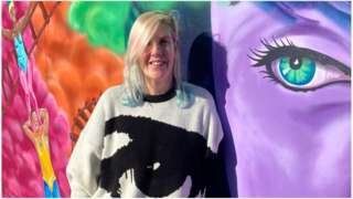 Street artist, Sarah Harris, stood against a wall displaying her artwork in Swindon. The painting includes a huge purple face with green eyes, a train track cutting through colourful clouds and the well known Swindon blondinie statue