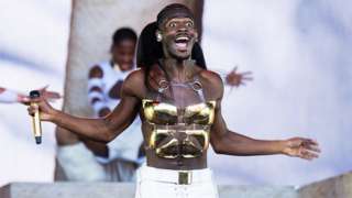 Lil Nas X has a beaming smile with his mouth open. A golden microphone is in his right hand, with both arms slightly outstretched. He is wearing a golden breast and stomach metal plate and white trousers. The background has a background dancer mid dance move, with a white and purple backdrop.