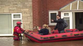 A couple and their dog being rescued in a dinghy boat from their flooded house