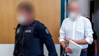 Bernd W, the German man convicted of murdering five people when he drove his car through a pedestrian zone, is led into court by a police officer. Both their faces are blurred out.