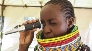 A Kenyan beekeeper holding a refractometer. She has her hair in cornrows and is wearing a traditional Kenyan clothing. She is squinting one eye.