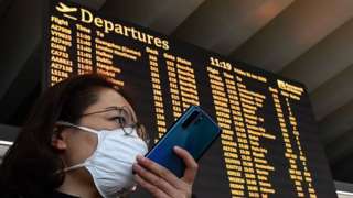 A passenger wearing a respiratory mask speaks on her smartphone by the departures board.