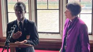 A Bishop in a purple robe and a young black school child speaking into a microphone in an ornate room