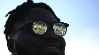 A man stands in the shade with bright reflections on his sunglasses.