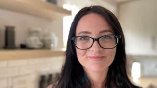 A woman with long, dark hair, wearing glasses is looking at the camera and smiling. Her kitchen is in the background.