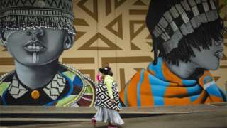 A mother carries her child on her back as she walks past graffiti artworks on the walls in the trendy Maboneng district of Johannesburg, South Africa, 02 October 2020. The area has been revitalized over the past five years as a process of re-development of the once downtrodden downtown district. The effort brought safety, business, more tourists and a lot of color into the area.