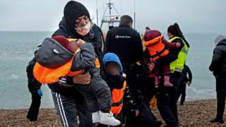 A migrant carries her children (with faces blurred) after being helped ashore from a RNLI (Royal National Lifeboat Institution) lifeboat at a beach in Dungeness, on the South East coast of England on 24 November 2021