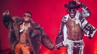 Wizkid and Burna boy performing on stage. Burna Boy is smiling and wearing a cowboy hat. Wizkid is wearing a big fur coat. Both have on sunglasses and jewellery. There is a red background.