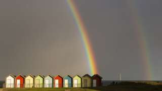 A rainbow archs across a dark sky and down on to a row of colourful beach huts