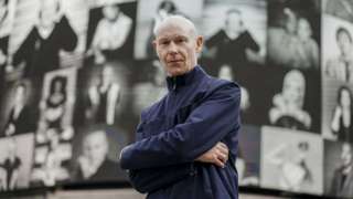 A portrait of a man with the backdrop of an exhibition behind him.