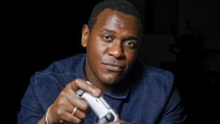 A close-up portrait against a black background of a man in a denim shirt holding a game controller, looking deep in concentration