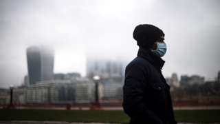 Man walks along south bank of Thames through city of London on a grey day, with a mask on