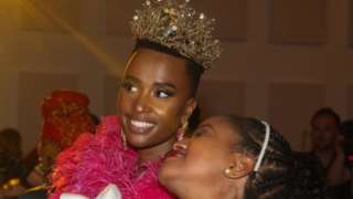 Two women at the "Bridgerton Affair" party smiling for a selfie. One of the women in wearing a crown and an elaborate pink dress. The other woman is dresses in a white outfit.