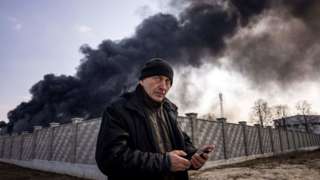 A man staring at the camera as he holds a phone with black billowing smoke in the background