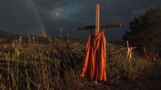 Crosses with dresses hung on them are seen by a road with a rainbow in the background