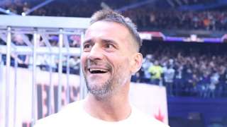CM Punk, a white man, at Survivor Series, wearing a white tshirt with his arm tattoos visible. He is looking at the crowd, with the background full of fans and flashing white lights.