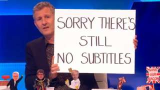 Adam Hills on Channel 4's The Last Leg holding up a sign reading "Sorry there's still no subtitles"