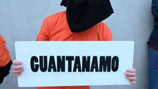 Hombre encapuchado con un cartel que dice Guantánamo