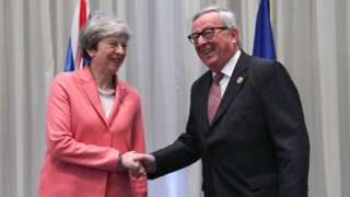 Theresa May and Jean-Claude Juncker smile and shake hands in front of the flags of their respective nations