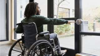 Stock image of a wheelchair user entering a building using a door access button