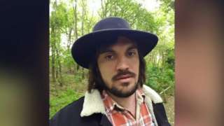Daniel Venes wearing a hat, checked shirt and coat in a woodland
