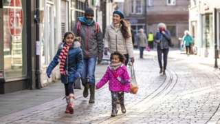 A family with two children walks down a high street wearing winter clothes