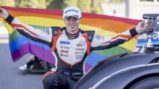 Richard Morris holding an LGBT flag by a trophy and racng car