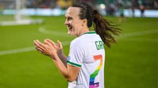 Ciara Grant in her Republic of Ireland kit which is white with green trim after a match in Dublin. She is clapping the fans and smiling. Her brunette hair is in a ponytail and the start of her name Grant is visible in green letters on the back of her shirt along with the number 2 which is in rainbow colours.
