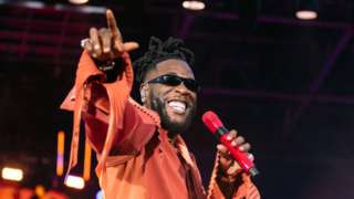 Burna Boy on stage at the London Stadium. He's wearing an orange jumpsuit and holding a red microphone in his left hand while smiling and pointing at the crowd with his right hand