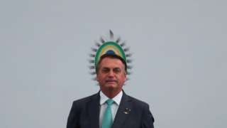 Bolsonaro em pé em frente a painel com símbolo brasileiro