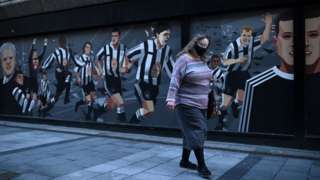 A woman wearing a mask walking past a Newcastle United mural in Newcastle