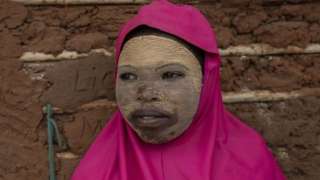 A girl wearing a clay mask and pink scarf smiles.